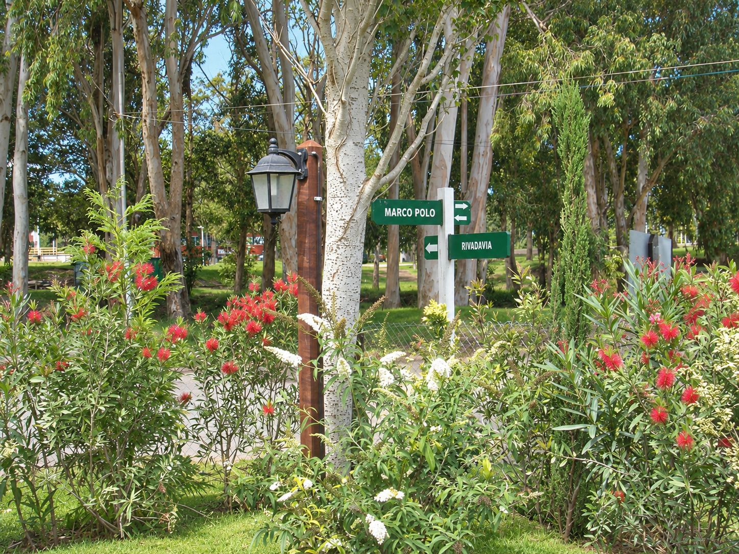 Jardín de la entrada de Monserrat Bungalows con flores rojas, un farol y carteles de calle entre los árboles